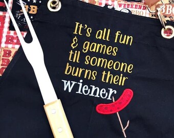 May include: Black barbecue apron with the text "It's all fun & games til someone burns their wiener" embroidered in yellow thread. A red and white hot dog is embroidered on the apron with a stick and two brown buns. A white grilling fork with a wooden handle is in the foreground.