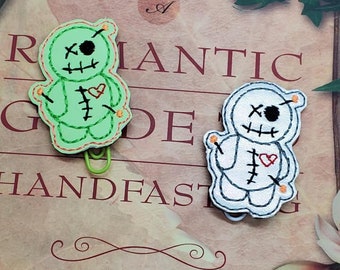 May include: Two green and white voodoo doll-shaped book marks with red hearts and stitched details. The book marks are on a book with the text "A Romantic Guide to Handfasting Rituals, Recipes & Lore".