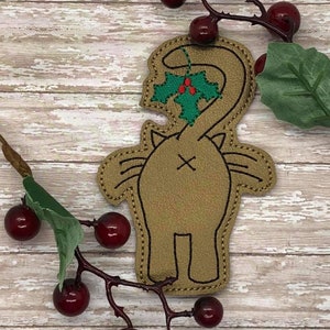 May include: A brown felt cat ornament with a green holly sprig and red berries coming out of its tail. The ornament is on a white wood background.