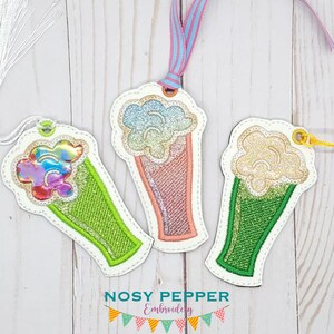Peut inclure: Trois marque-pages brodés colorés en forme de cornets de glace. Chaque marque-page a un schéma de couleurs différent et une texture scintillante. Les marque-pages sont sur un fond en bois blanc.