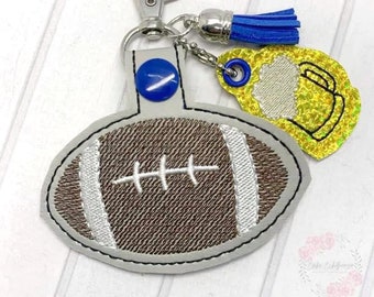 Puede incluir: Un llavero con forma de balón de fútbol marrón y blanco con un borla azul y un dije de jarra de cerveza amarilla. El llavero está unido a un llavero plateado.