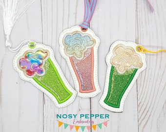 May include: Three colorful embroidered bookmarks shaped like ice cream cones. Each bookmark has a different color scheme and a glittery texture. The bookmarks are on a white wooden background.