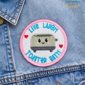 Könnte beinhalten: Gestickter Aufnäher mit rosa Rand und den Worten "Live Laugh Toaster Bath" in Blau. Der Aufnäher zeigt einen lächelnden Toaster mit Herzen und ist an einer Jeansjacke befestigt.