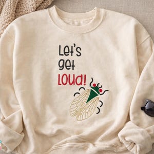May include: Cream-colored sweatshirt with the phrase "Let's get loud!" in black and red text. A green and tan insect graphic is below the text. Black sunglasses and a coffee cup are in the image.