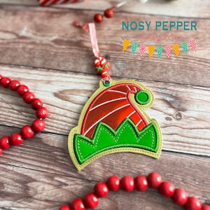 May include: A gold, green, and red felt elf hat ornament with a candy cane and a green bead hanging from it. The ornament is on a wooden surface with red beads.