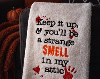 May include: White Halloween-themed towel with the text "Keep it up, & you'll be a strange SMELL in my attic" in black and orange. Red blood splatter and handprints add to the spooky design. Perfect for Halloween decor.
