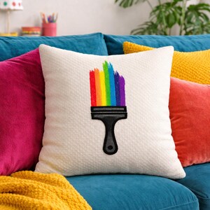 Könnte beinhalten: Ein weißes Zierkissen mit einem Regenbogen-Pinsel-Design. Der Pinsel hat einen schwarzen Griff und Borsten in Rot, Orange, Gelb, Grün, Blau und Lila. Das Kissen liegt auf einem blauen Sofa mit anderen bunten Kissen.