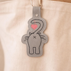 Op de afbeelding: Een grijze katvormige sleutelhanger met een roze hart en zwarte omtrekdetails. De sleutelhanger is bevestigd aan een canvas draagtas. Het ontwerp bevat de achterkant en de staart van een kat. De sleutelhanger is gemaakt van vilt en heeft een decoratieve strass-steen.