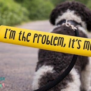 Może przedstawiać: Żółty uchwyt do smyczy dla psa z czarnym napisem "I'm the problem, it's me". Uchwyt jest przymocowany do czarnej smyczy, z czarno-białym psem w tle.