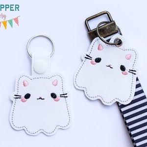 May include: Two white ghost-shaped keychains with embroidered cat faces. The cat faces have pink ears, black eyes, and pink cheeks. One keychain is attached to a silver key ring and the other is attached to a black and white striped ribbon.