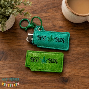 May include: Two rectangular lighter sleeves made of green glitter fabric. One sleeve features a green metal keyring and holds a silver lighter. Both sleeves are embroidered with the black text BEST BUDS and a central green cannabis leaf icon.