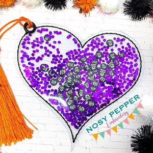 May include: A clear heart-shaped bookmark with a purple glitter and black skull design. The bookmark has a black tassel and the text "NOSY PEPPER Embroidery" is printed on the bottom.