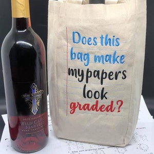Może przedstawiać: Biała płócienna torba na wino z haftowanym wzorem w kolorze niebieskim i czarnym, na którym widnieje napis "Does this bag make my papers look graded?" Torba leży na stole obok butelki wina.