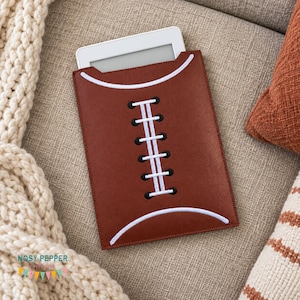 Puede incluir: Una funda rectangular marrón para lector electrónico diseñada para parecer un balón de fútbol americano. La funda presenta líneas blancas curvas en la parte superior e inferior, con un patrón de cordones blancos verticales cosido sobre ojales negros.