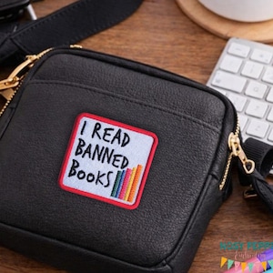 May include: Black crossbody bag with a gold zip and a patch reading "I READ BANNED BOOKS." The patch has a red border, white background, and a stack of colourful books. The bag features a black strap.