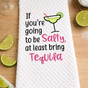 May include: White kitchen towel with a textured pattern, embroidered with the phrase "If you're going to be Salty, at least bring Tequila." A lime margarita graphic is also included. Limes and salt are in the background.