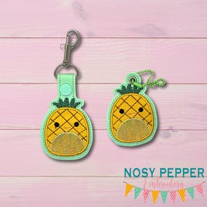 Op de afbeelding: Twee geborduurde ananas sleutelhangers met een schattig cartoon gezicht. De ananas is geel met een groene bovenkant en bevindt zich op een lichtgroene achtergrond. De sleutelhangers zijn gemaakt door Nosy Pepper Embroidery.
