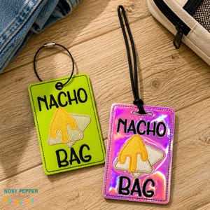May include: Two rectangular luggage tags with the words "NACHO BAG" in black. One tag is lime green, the other is iridescent pink. Each tag features an embroidered nacho chip design with yellow cheese. A black loop and strap are attached.