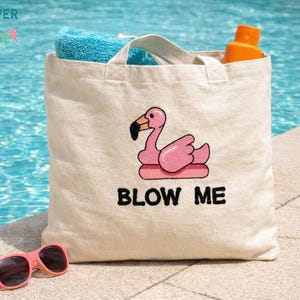 May include: A beige tote bag with a pink flamingo design and the text "BLOW ME" in black. The bag contains a blue towel and an orange bottle. Pink sunglasses rest in front of the bag.