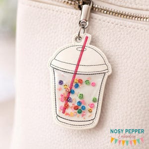 May include: A white keychain charm shaped like a beverage cup, featuring a red straw and filled with colorful smiley face beads. The charm is attached to a silver metal clasp.
