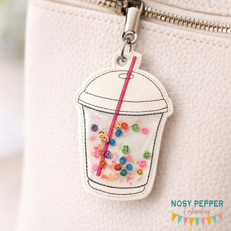 May include: A white keychain charm shaped like a beverage cup, featuring a red straw and filled with colorful smiley face beads. The charm is attached to a silver metal clasp.