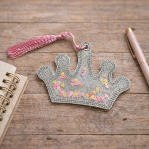 May include: A silver crown-shaped bookmark with colorful confetti and a pink tassel. A spiral notebook and a pink pen with a gold tip are also on the wooden surface. The bookmark is a decorative item.