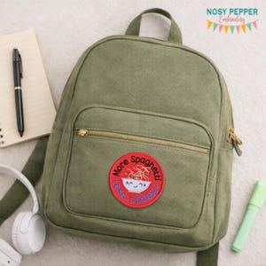 May include: Olive green backpack with a front zippered pocket and a top handle. A round patch with the text "More Spaghetti Less Upsetti" and a bowl of spaghetti is sewn on the front. A pen, notepad, headphones, and highlighter are in the background.