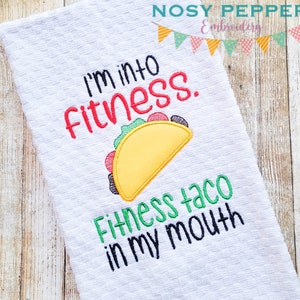 以下が含まれることがあります： 白いキッチンクロスに「I'm into fitness. Fitness taco in my mouth」という刺繍が施され、黄色のタコスに緑のレタス、赤いトマト、茶色の肉が乗っています。
