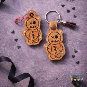 May include: Two brown voodoo doll keychains. Each keychain features a stitched design with an "X" for eyes, a heart, and stitched details. One has a red beaded chain, the other a metal ring with a brown tassel. The background is a purple surface with black heart shapes.