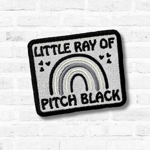 Pode incluir: Patch bordado com o texto "LITTLE RAY OF PITCH BLACK" acima de um design de arco-íris em tons de cinza. O patch tem uma borda preta e pequenos corações. O patch quadrado parece ser feito de tecido.