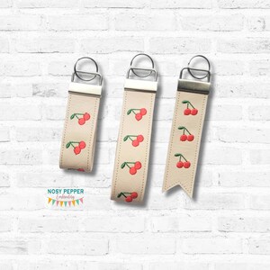 May include: Three cream-colored keychains with embroidered red cherries and green leaves. Each keychain has a silver-colored metal ring and a cream-colored leather strap. The text "NOSY PEPPER Embroidery" is at the bottom.