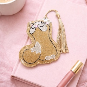 Op de afbeelding: Een gouden glitter bladwijzer in de vorm van een kat, met een gouden kwast, ligt op een roze boek. De bladwijzer heeft een witte buik en gesloten ogen. Een kopje koffie, zonnebril en lipgloss staan ook in de afbeelding.