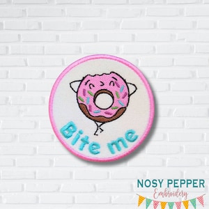 May include: Pink and brown embroidered donut with sprinkles and a smiling face. The donut is holding a sign that says "Bite me".