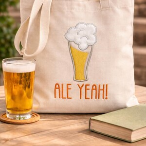 Könnte beinhalten: Eine cremefarbene Canvas-Tragetasche mit einem Bierglas-Design und dem Text "ALE YEAH!". Ein Bierglas steht auf einem Holztisch neben der Tasche. Ein Buch und ein Untersetzer befinden sich ebenfalls auf dem Tisch.