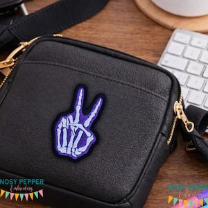 May include: Black crossbody bag with a gold zipper and a purple and white embroidered peace sign patch. The patch features a skeleton hand in a peace sign gesture. The bag has a black strap and a gold zipper.