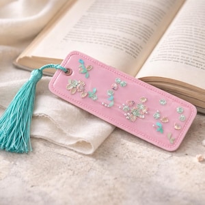 May include: A pink bookmark with floral and butterfly embellishments and a turquoise tassel rests on a book. The bookmark has a rectangular shape with rounded corners and a small hole at the top for the tassel.