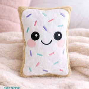 May include: A plush, rectangular pillow shaped like a slice of toast, with a smiling face and colorful sprinkles. The pillow has a white center with a tan border, and black eyes and a curved mouth. The sprinkles are pink, purple, and blue.