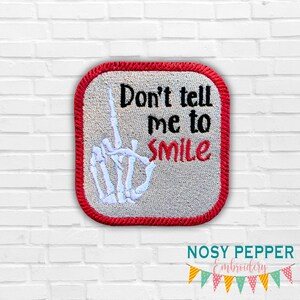 May include: A square embroidered patch with a red border. The patch features a white skeleton hand giving the middle finger and the text "Don't tell me to smile" in black and red.