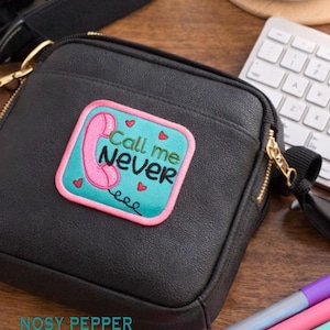 Peut inclure: Sac bandoulière noir avec une fermeture éclair dorée et un écusson « Call me Never » avec un téléphone rose. L'écusson est sur un fond turquoise avec des cœurs rouges.