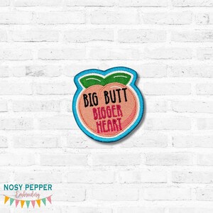 Peut inclure: Écusson brodé en forme de pêche avec les mots "BIG BUTT BIGGER HEART" en noir et rouge. La pêche est de couleur pêche avec des feuilles vertes et une bordure bleue. L'écusson est sur un mur de briques blanches.