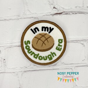 May include: A round, white embroidered patch with a brown sourdough bread design and the text "In my sourdough era" in green and black lettering.