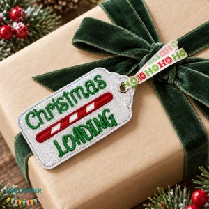 May include: A silver glitter gift tag with green embroidered text that reads Christmas Loading and a red and white candy cane graphic. The tag is attached with a ribbon featuring repeating red and green Ho Ho Ho text to a brown gift box with a dark green velvet bow.