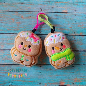 May include: Two gingerbread man plush toys with embroidered details, one with a green scarf and the other with a pink bow, attached to a green and pink plastic keychain.
