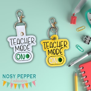 以下が含まれることがあります： 黒い文字で「Teacher Mode On」と書かれたキーホルダーが2つ。1つは緑色の「オン」スイッチが付いた白いキーホルダー、もう1つは緑色の「オン」スイッチが付いた黄色のキーホルダーです。