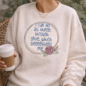 Op de afbeelding: Crèmekleurige sweatshirt met de tekst "I can do all things through spite, which strengthens me" in een cirkelvormig ontwerp. Een paarse roos en groene bladeren staan onder de tekst. De sweatshirt is een casual stijl.