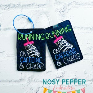 May include: Two black luggage tags with a blue loop. The tags have white text that reads "Running on Caffeine & Chaos". The text is surrounded by a graphic of a skeleton hand holding a pink and green beverage.