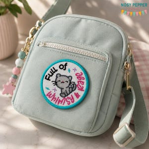 May include: A light sage green crossbody bag with a front zippered pocket and an adjustable strap. A circular embroidered patch features a gray cat illustration with the text "Full of Whimsy & Rage". A gold chain with pastel beads hangs from the side.