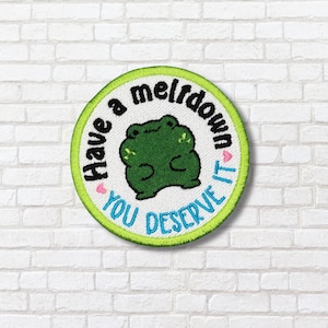 Puede incluir: Parche bordado redondo con borde verde y el texto "Have a meltdown, you deserve it". Una ilustración de rana verde está en el centro. El parche está sobre una pared de ladrillos blancos.