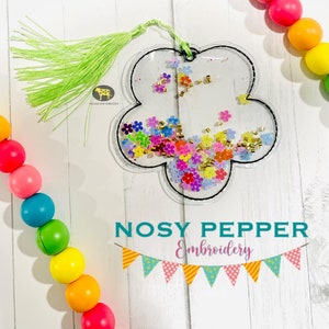 May include: A clear plastic flower-shaped bookmark with a green tassel. The bookmark is filled with colorful glitter and small gold beads. The bookmark has a black outline and the text "NOSY PEPPER Embroidery" is printed below it.