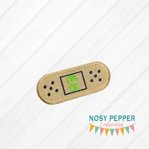 Puede incluir: Un parche de planchado de color beige con forma de vendaje, bordado con un cuadrado negro que contiene el mensaje verde "I'm OK".